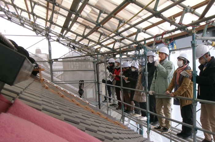 工事現場の足場をのぼり、屋根を間近に見ながら解説する職員と解説を聞く参加者の写真