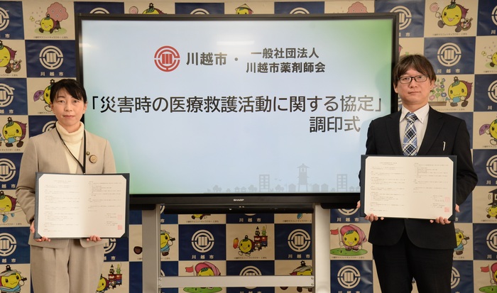 協定調印式の写真。「災害時の医療救護活動に関する協定」と書かれたモニターの前に、市長と川越市薬剤師会の会長がそれぞれ協定書を持ち立っている。